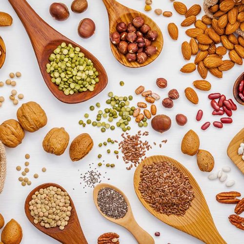 Various legumes and different kinds of nuts in spoons. Walnuts kernels ,hazelnuts, almond ,brown pinto ,soy beans ,flax seeds ,chia ,red kidney beans and pecan set up on white wooden table.