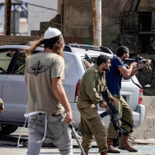 Israeli settlers fire at Palestinians (unseen) while an Israeli soldier (L) stands by during clashes in the town of Huwara in the occupied West Bank on October 13, 2022. (Photo by Oren ZIV / AFP)