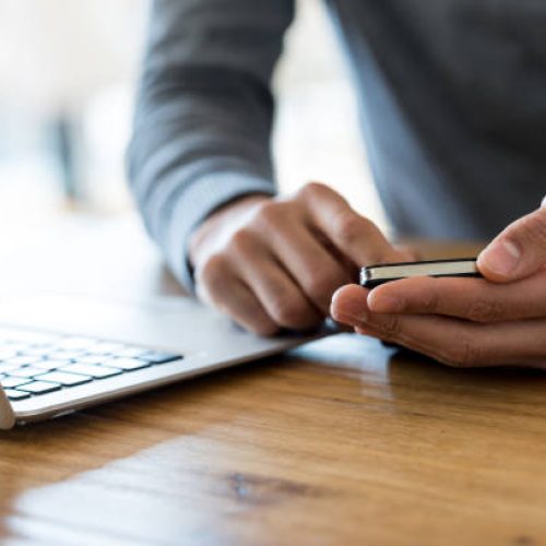 Close-up of unrecognizable man in grey sweater sitting at wooden table next to laptop and holding cell phone in both hands