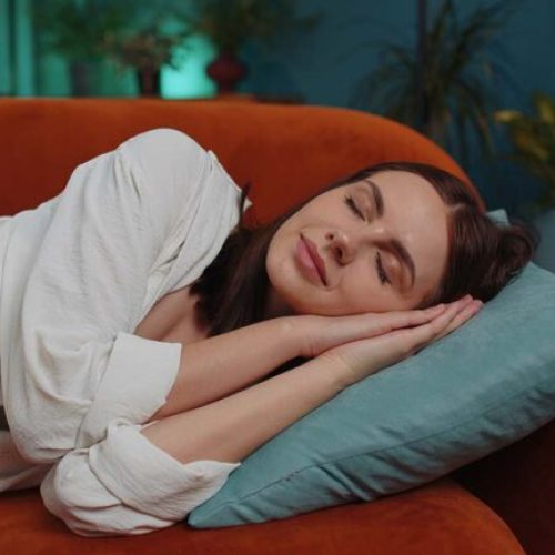 Tired caucasian adult girl lying down in bed taking a rest at home. Carefree young woman napping, falling asleep on comfortable orange sofa with pillows. Closed her eyes enjoy daytime nap alone
